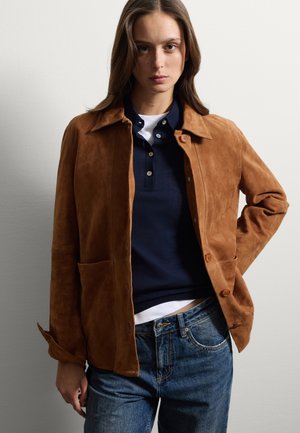 Giacca in suede marrone con colletto classico, chiusura frontale con bottoni e due grandi tasche. Indossata sopra un polo blu navy e jeans.