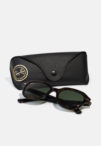 Occhiali da sole con lenti verde scuro in una montatura tartarugata. Include un astuccio nero testurizzato con un logo dorato e chiusura a scatto.