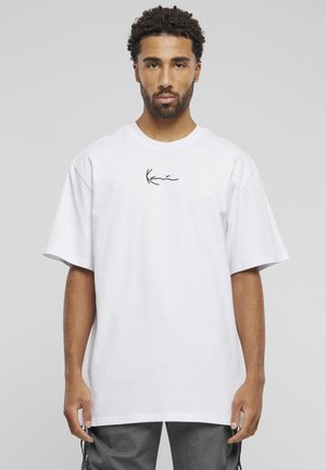 Homme aux cheveux courts et bouclés portant un t-shirt blanc uni avec un petit texte noir de signature sur la poitrine, debout devant un fond gris.