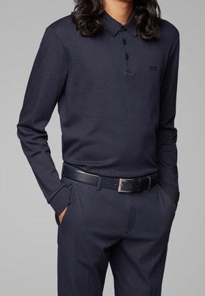 Homme portant un polo bleu marine à manches longues et un pantalon assorti avec une ceinture en cuir noire, mains dans les poches, sur fond gris uni.