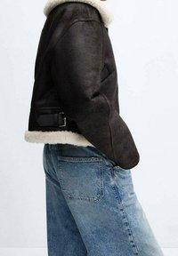 Sort faux læderjakke med en hvid shearling krave og manchetter; har et sidebuckle-detajle og en tætsiddende silhuet. Båret med blå jeans.