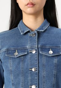 Jeans jakna u srednje plavoj boji s klasičnim ovratnikom, srebrnim kopčama i dva džepa na prsima. Tkanina ima mekan i teksturiran izgled.