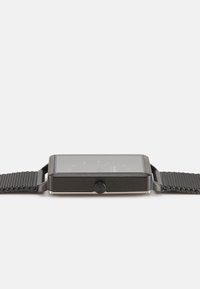 Montre carrée avec un boîtier en métal noir et un bracelet en mesh sombre. Présente un cadran en verre clair et des marqueurs d'heure minimalistes. Bouton sur le côté.