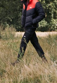 Giacca a vessillo nera e rossa con colletto alto, abbinata a pantaloni cargo neri. L'outfit presenta un logo sul petto e sui pantaloni. Ambientazione all'aperto.