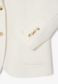 Blazer bianco sporco con maniche e parte frontale, bottoni beige e logo ricamato discreto vicino al polsino su tessuto testurizzato.