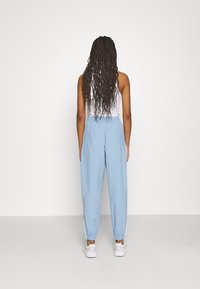 Kvinna med långt flätat hår, iklädd en vit linne, ljusblå joggingsbyxor och vita sneakers, som står framför en helt vit bakgrund.