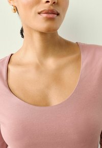 Hellrosa Scoop-Neck-Top aus weichem Stoff, mit glatter Textur und subtiler Schattierung. Begleitet von runden goldenen Ohrringen.