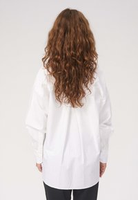 Hvid skjorte med en afslappet pasform, lange ærmer, afrundet kant og knaplukninger ved ærmerne. Stoffet ser glat ud med en subtil glans.