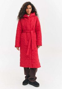 Czerwony pikowany długi płaszcz z kapturem, z fakturą w liście, z paskiem w talii, bocznymi kieszeniami, noszony na brązowych spodniach i czarnych kapciach.