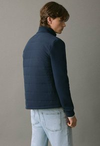 Mlađi muškarac u tamnoplavoj jakni s rombastim uzorkom i pletenim rukavima, okrenut bokom prema jednostavnoj sivoj pozadini.