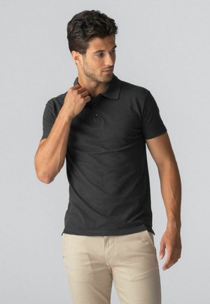 Mees reguleerib tumehalli polo-särgi kraed, seljas beežid püksid, seisab tavalise heleda taustaga.