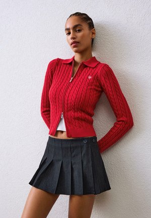 Rød kabelstrikket cardigan med krave og lynlås, parret med en sort plisseret nederdel med striber og knapdetaljer.