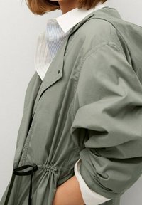 Lichtgroene, lichtgewicht jas met een verstelbare aansnoerbare taille, oversized mouwen en een capuchon, gedragen over een gestreept overhemd met kraag.