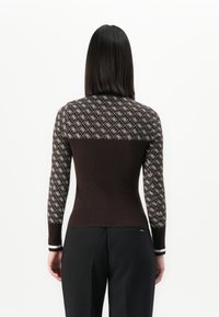 Mujer de espaldas que lleva un suéter ajustado marrón con un patrón de diamantes blancos en la parte superior de la espalda y las mangas, combinado con pantalones negros.