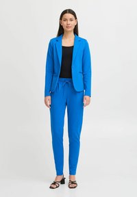 Modrý blazer a odpovídající kalhoty vyrobené z hladkého materiálu. Blazer má jeden knoflík, kalhoty mají pás se šňůrkou. Černý top pod tím.