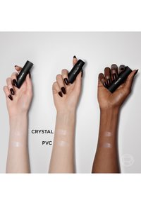Drie handen houdt zwarte cilindrische cosmetische verpakkingen met een witte bovenkant vast, met licht en donker huidtinten van producten gelabeld als "CRYSTAL" en "PVC."