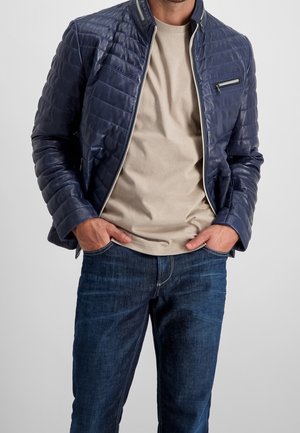 Mann trägt eine blaue gesteppte Jacke über einem beigen T-Shirt, mit den Händen in den Taschen und dunkelblauen Jeans vor einem einfarbigen Hintergrund.