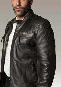 Veste de moto en cuir marron avec des détails de quiltage en losange sur les épaules, poches zippées à l'avant et logo sur la poitrine. Texture lisse.