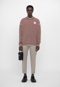 Braun-weiß gestreifter Pullover mit Rundhalsausschnitt und kleinem Logo, kombiniert mit beigen Hosen und schwarzen Sneakern, während eine schwarze Tasche gehalten wird.
