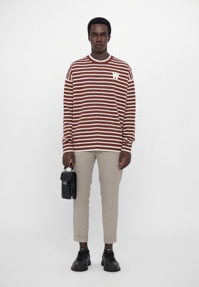 Suéter a rayas marrón y blanco con cuello redondo y pequeño logo, combinado con pantalones beige y zapatillas negras, sosteniendo un bolso negro.