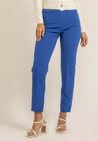 Pantalons ajustés bleu fabriqués à partir d'un tissu extensible avec une texture lisse. Comprend des poches avant et une taille plate. Associés à des sandales blanches.