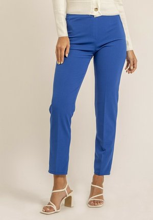 Pantalons ajustés bleu fabriqués à partir d'un tissu extensible avec une texture lisse. Comprend des poches avant et une taille plate. Associés à des sandales blanches.