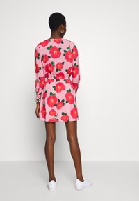 Rosa Blumen-Kleid mit langen Ärmeln, geraffter Taille und glockenförmigem Rock, verziert mit roten Blumen und grünen Blättern. Getragen mit weißen Turnschuhen.