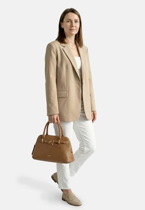 Donna con blazer beige e pantaloni bianchi che tiene una borsa in pelle marrone, in piedi, guarda avanti con espressione neutra.