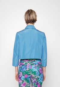 Giacca blu tagliata con colletto e maniche lunghe, realizzata in tessuto liscio. Indossata con pantaloni floreali a fantasia vivace, caratterizzati da tonalità di rosa e verde.