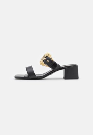 Sandalia negra de cuero con tacón y dos tiras, con una hebilla decorativa dorada con la etiqueta "COUTURE" en la tira superior.