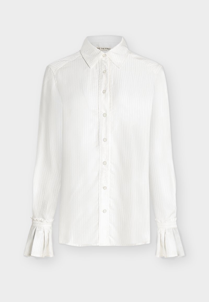 Λευκό μπλουζάκι με μακριά μανίκια, κουμπιά στο μπροστινό μέρος, μυτερός γιακάς, διακριτικές κάθετες ρίγες και φαρδιά μανσέτα με λεπτομέρειες βολάν.