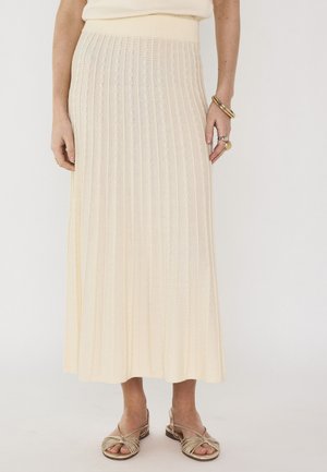 Femme portant une jupe midi plissée crème, des sandales dorées et des bijoux en or aux mains, sur un fond clair uni.