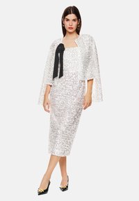 Weißes Pailletten-Kleid mit geradem Schnitt und knielangem Saum, kombiniert mit einer transparenten, kurzärmligen Cape und einem schwarzen Ripsband-Detail. Schwarze Absätze vervollständigen den Look.