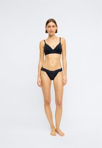 Schwarzes Bralette mit V-Ausschnitt und dünnen Trägern, kombiniert mit passenden schwarzen Bikini-Unterhosen mit Spitze. Glatte Textur, minimalistischer Stil.