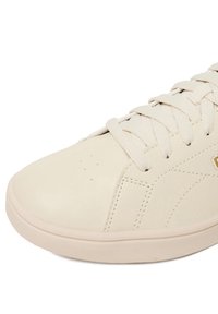 Detail béžovej špičky tenisky s textúrovanou podrážkou, prešitými časťami a zladenými šnúrkami na bielom pozadí.