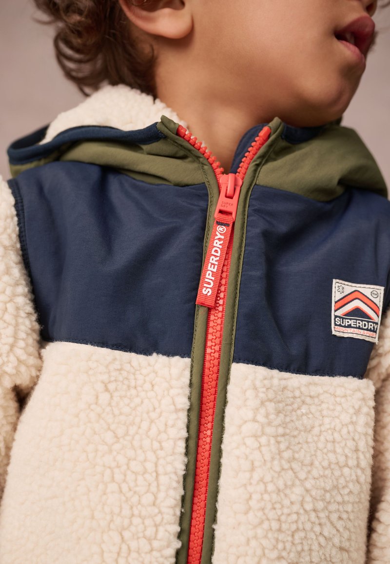 Fleecová bunda s kapucňou v námorníckej modrej hornej časti, krémovej dolnej časti a zelenými akcentmi; obsahuje výrazný červený zips a nášivku s logom na hrudi.