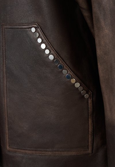 Veste en cuir brun avec une poche latérale, ornée de clous métalliques sur le bord. Texture lisse avec un léger éclat et des coutures détaillées.