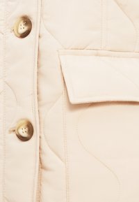Ανοιχτό μπεζ μπουφάν με κοντές ραφές που διαθέτει έντονη κούμπωμα με κουμπιά και ορθογώνια τσέπη με κυρτές λεπτομέρειες ραφής. Απαλή υφή.