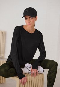Schwarzes Langarmoberteil mit Daumenlöchern, grüne Tarnleggings und weiße Sneakers. Das Model trägt eine schwarze Kappe mit einem geprägten Logo.
