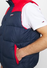 Tmavě modrá prošívaná vesta s červenou kapucí a rameny, vybavená předním zapínáním na knoflíky a bočními kapsami s červeným lemováním.