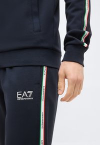 Σκούρο μπλε φόρμα με λευκό λογότυπο EA7 Emporio Armani, ρίγα σε κόκκινο, λευκό και πράσινο στο μανίκι και στο πόδι του παντελονιού, το χέρι χαλαρό στο πλάι.