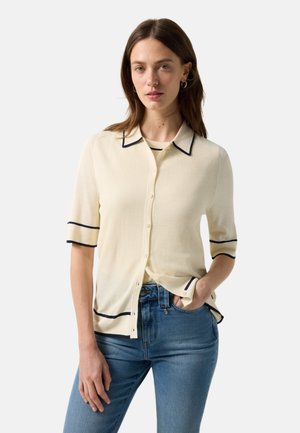 Kobieta w beżowym, zapinanym na guziki, krótkim sweterku z czarną lamówką, nosząca beżowy top i niebieskie dżinsy, stoi z jedną ręką w kieszeni.