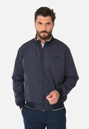 Uomo con barba che indossa una giacca bomber blu navy con cerniera e pantaloni scuri, in piedi contro uno sfondo chiaro uniforme.