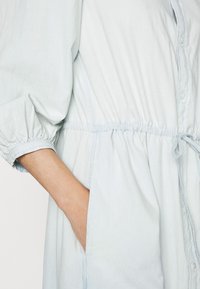 Vestito blu chiaro con bottoni, a vita stretta e maniche a palloncino. Realizzato in un tessuto morbido e leggero. Presenta tasche laterali e un cordoncino.