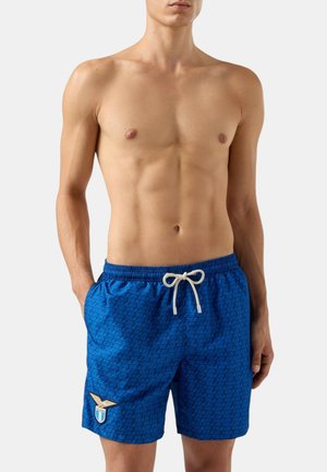 Uomo senza maglietta che indossa pantaloncini da bagno blu con coulisse bianca e un piccolo emblema sulla gamba sinistra, in piedi davanti a uno sfondo semplice.