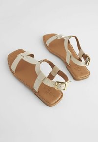 Beige platta sandaler med korsande remmar, med fyrkantig tå och spänne. Slät yta med brun innersula och sömnadsdetalj.