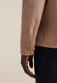 Manica di giacca marrone chiaro con due bottoni decorativi. Il tessuto appare liscio con cuciture visibili. Mano parzialmente visibile, indossando pantaloni scuri.