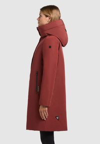 Parka imperméable rouge avec un col haut et une grande capuche, comportant une poche zippée sur le côté et un logo triangulaire sur l'ourlet.