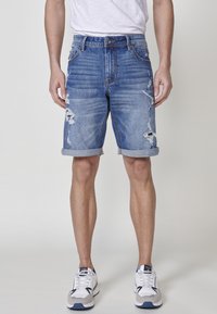 Denim šortsevi u svijetloplavoj boji s iznošenim detaljima, prebacenim manžetama i klasičnim dizajnom s pet džepova, noseni s bijelim i sivim tenisicama.