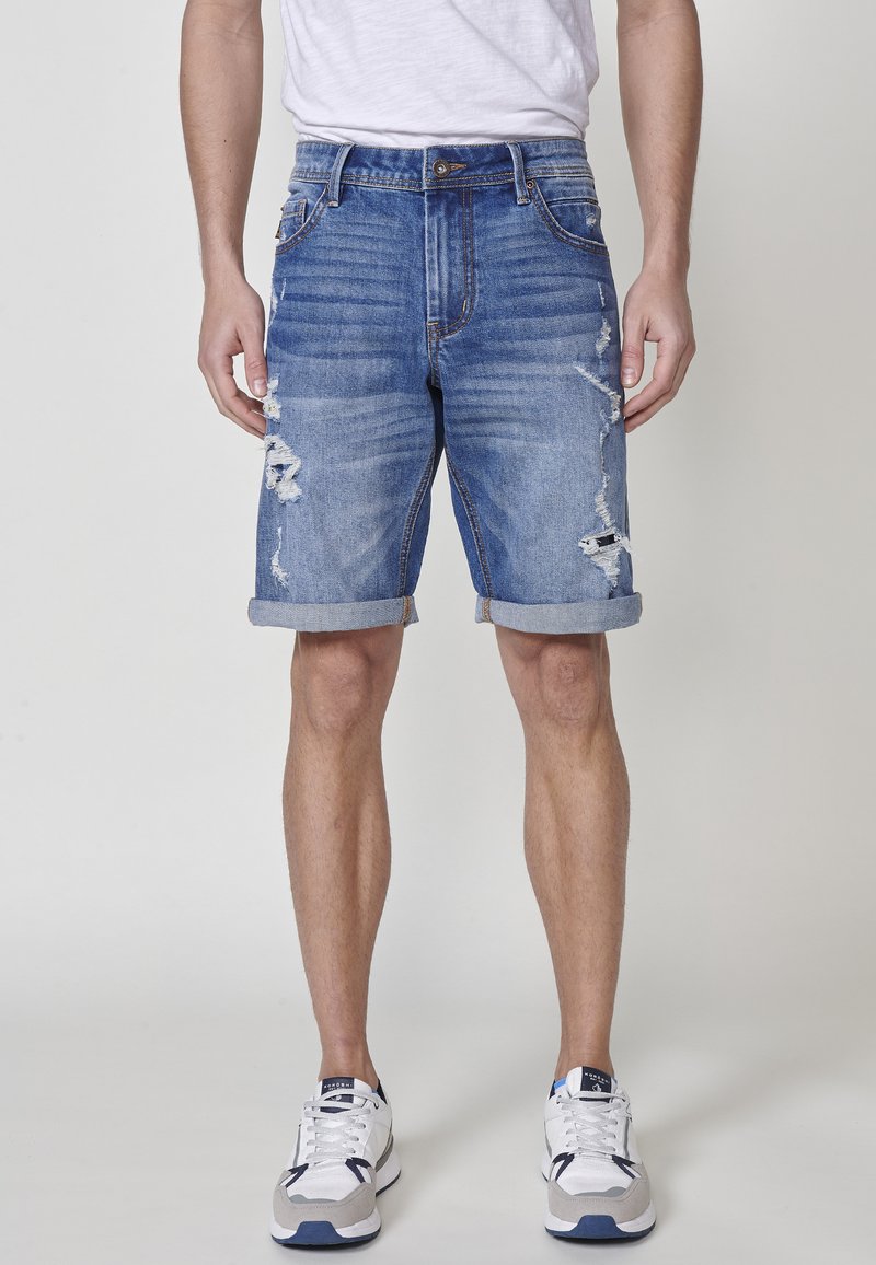 Denim šortsevi u svijetloplavoj boji s iznošenim detaljima, prebacenim manžetama i klasičnim dizajnom s pet džepova, noseni s bijelim i sivim tenisicama.